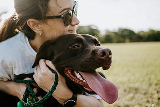 A very dark brown dog being hugged by a woman with sunglasses. The background appears to be an open park or exterior.