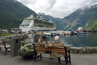 A retired couple sitting on a bench watching a cruise ship by the mountains.