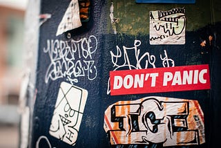 Stickers and graffiti on a wall. One says, “Don’t Panic.”