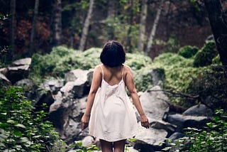 A Black woman, her back to the camera, wearing a white slip dress, stands in the middle of a forest, with beautiful trees and white flowers scattered across the ground. Her arms a stretched out and she is looking up.