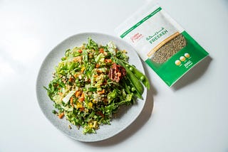 a bag of freekeh seeds next to a plate of salad with veggies and grain, the grain is freekeh.