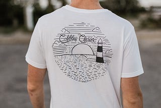 White t shirt with a black graphic on top. Picture of a subset and lighthouse