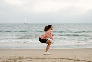 Woman in a pink sweater and black shorts doing yoga on a beach