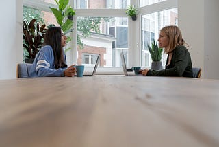 Two women with laptops facing each other at a conference room table.