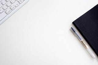 A keyboard, planner and pen placed on a table top.