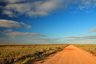 A red dirt road stretches into the horizon of the Nullarbor Plain under a vast sky with scattered clouds.