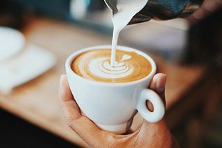 Image shows someone’s hand holding a coffee cup, while milk is being poured into creating a perfect looking coffee.