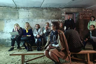 A group of people wearing coats and scarves and sitting on benches while holding a meeting in an underground bunker in Ukraine.