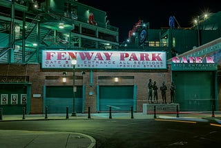 A night view of Fenway Park’s entrance with its iconic red and white signage. The image shows the “FENWAY PARK” sign above Gate B, indicating entrances for Van Ness Street and Ipswich Street. To the right, a “Gate K” sign marks the kids’ entrance. A statue of Ted Williams and other players stands in front, bathed in the soft glow of streetlights, creating a nostalgic and peaceful atmosphere outside the historic ballpark.