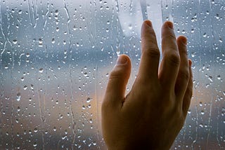 A person’s hand rests against the inside of a window that’s fogged and splattered with rain drops with a view upon an ambiguous landscape of diffused golden colors near the bottom and blue-grey colors near the top, casting an impression of a damp, cloudy morning.