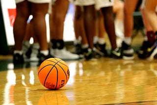 A basketball sits on a school gym floor among an ankle shot of girls in bym shorts.
