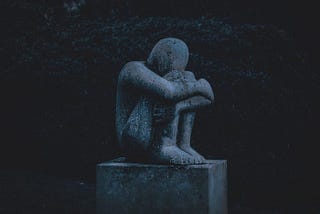 Sculpture of boy sits on a pillar, knees against his forehead, arms hug his folded up legs.