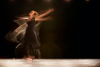 Uma dançarina, fazendo seus movimentos fluidos em uma imagem que evidencia o contraste da iluminação direta e focada na dançarina com um fundo escuro.