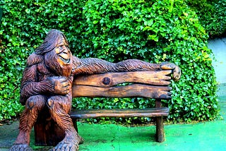 Bigfoot statue sitting on the left side of a bench looking to his right.