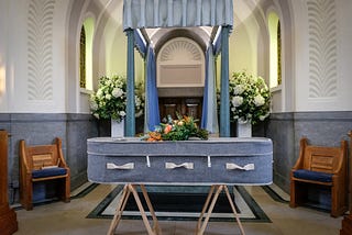 Photo of a blue-grey casket on wooden sawhorses, in front of a blue, curtained archway, set up in an alcove. There are flowers on top of the casket and behind the curtained structure. The walls behind are white with ornate designs on the upper part of the wall, and blue-grey on the lower part of the walls to the wainscotting. There’s a small, wooden bench with blue, padded seat on either side of the image.