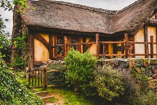 A set of stairs cut into the earth lead up to a wood and plaster cottage with a thatched roof. A stone wall surrounds part of the perimeter, and various bushes and shrubs grow near the wall.