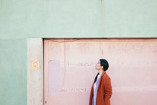 A woman in a fall coat stands outside a pastel-painted concrete building looking to the sky as though she is thinking.