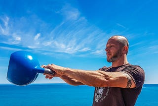 A man helps maintain his weight by swinging  a blue kettlebell. The ocean is in the background.