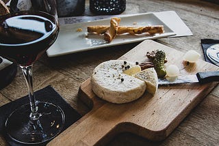 Dark red wine in a tall wine flute on a black napkin next to a wooden cutting board with a cheese wheel, a pie slice cut out, adjacent to a knive and bits of herb and cheese. Behind the cutting board is a ceramic tray with a twisted bread stick on it.