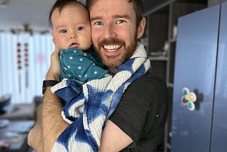 An image of the author holding his infant daughter. Both are looking at the camera. The infant is wearing a teal onesie with white polka dots and has a neutral gaze. The author is wearing a short-sleeved dark tee shirt and has both of his arms wrapped around his daughter. He is smiling widely.