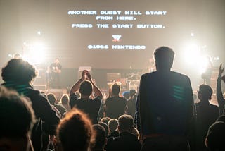 Facing a stage with a rock band on it, three people stand to clap while many more are seated. On the screne above the performers are the words, “Another Quest will start from here Press the start button.”