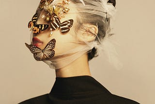 A portrait photo of a light-skinned woman with white gauzy fabric covering her face and five butterflies perched on top of the fabric, obscuring her eyes and nose.