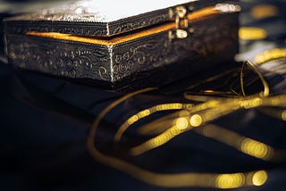In this picture, a small antique-looking chest is displayed, glowing with a golden light from within. The chest is a light silver-grey color and is surrounded by golden coins.