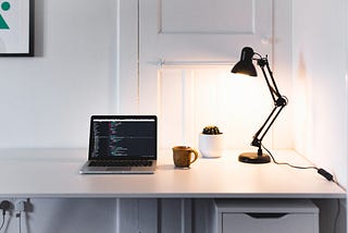 Laptop on top of a white desk, with code on the screen, next to an orange mug, a cactus and a black lamp