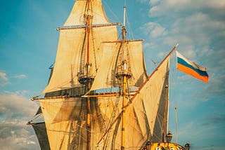 A classic tall ship or galleon with multiple sails, sailing at sunset with a flag featuring horizontal stripes