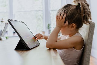 A six-to-eight-year-old child uses her iPad while her arm rests on the table and her hand rests on her forehead.