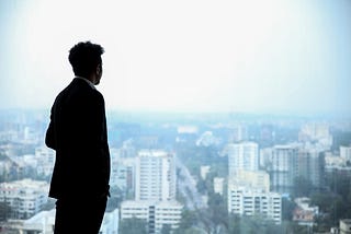 Man in dark suit looking out of corporate office window over panorama of other corporate office buildings