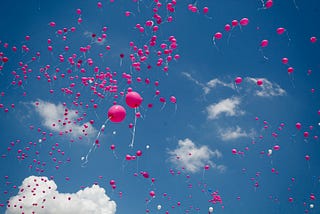 Many pink balloons flying towards a very blue sky, with some clouds.