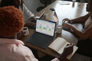 A group of three people sitting around a wooden table with laptops. One person is working on a laptop displaying a calendar or scheduling tool in an Excel-like interface. The individual is holding a coffee cup while taking notes. This image illustrates a collaborative working environment where team members are discussing or reviewing data on their laptops.