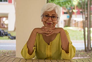 Asian woman with white hair and glasses, hands folded under her chin, wearing a bright yellow shirt