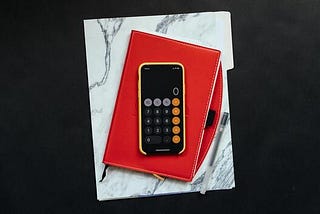 A mobile phone has the calculator app open which sits on top of a red notebook and white marble folder.