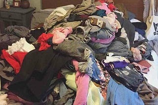 A mountain of clothes, mostly blacks, greys, and pinks, piled atop the author’s bed.