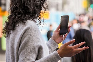Exasperated lady in gray sweater, long curly black hair, head away from camera, holding phone in left hand, and right hand extended