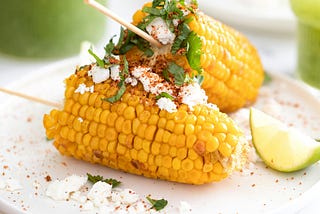 corn on the cob broken in half then put on a skewer for easy eating, served with a sprinkle of cheese, spices, fresh herbs and a wedge of lime on a white plate