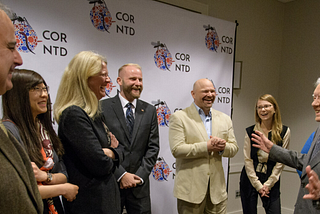 Six representatives from USAID stand with President Jimmy Carter. USAID employees smile and laugh as he makes a joke and spreads his arms out in front of him.