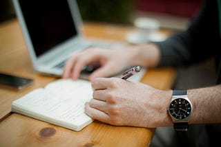 Desk, laptop, notebook open, pen in left hand, right hand resting