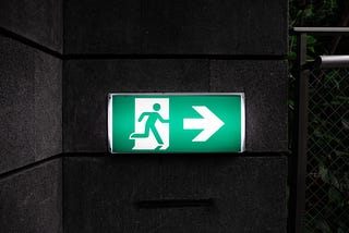 Image of a green and white illuminated exit sign on a dark background. The sign shows the image of a person leaving through a doorway, and has an arrow pointing to the right.