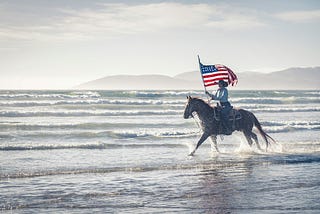 Man on horse galloping through waves while holding an American flag.