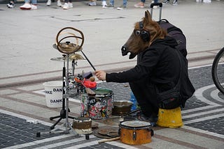 A man in a costume of a horses head, playing drums on the road