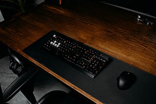 A Black Keyboard on a wooden desk with a monitor in sleep mode which says “DO MORE” across the screen in large white text