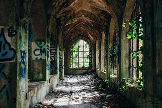 A crumbling stone corridor with arched windows. There is graffiti on some of the walls and ivy breaking through the windows on the right.