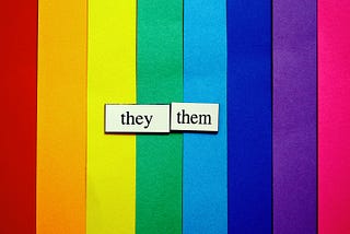 [image description: vertical strips of paper that form a rainbow and a slip of white paper that says “they/them” in black.]