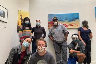 Seven figures standing in front of art on walls, all wearing face masks.