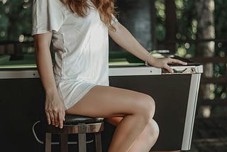 A hot free-use red head sits next to a billiards table on a stool.
