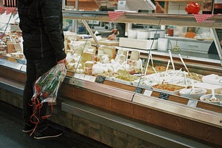 A person in a black outfit stands in front of a brightly lit deli counter, which holds cheeses, and several white bowls of various deli foods.