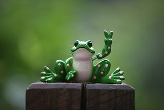Figurine of a frog cheerily holding up the two-finger peace sign.